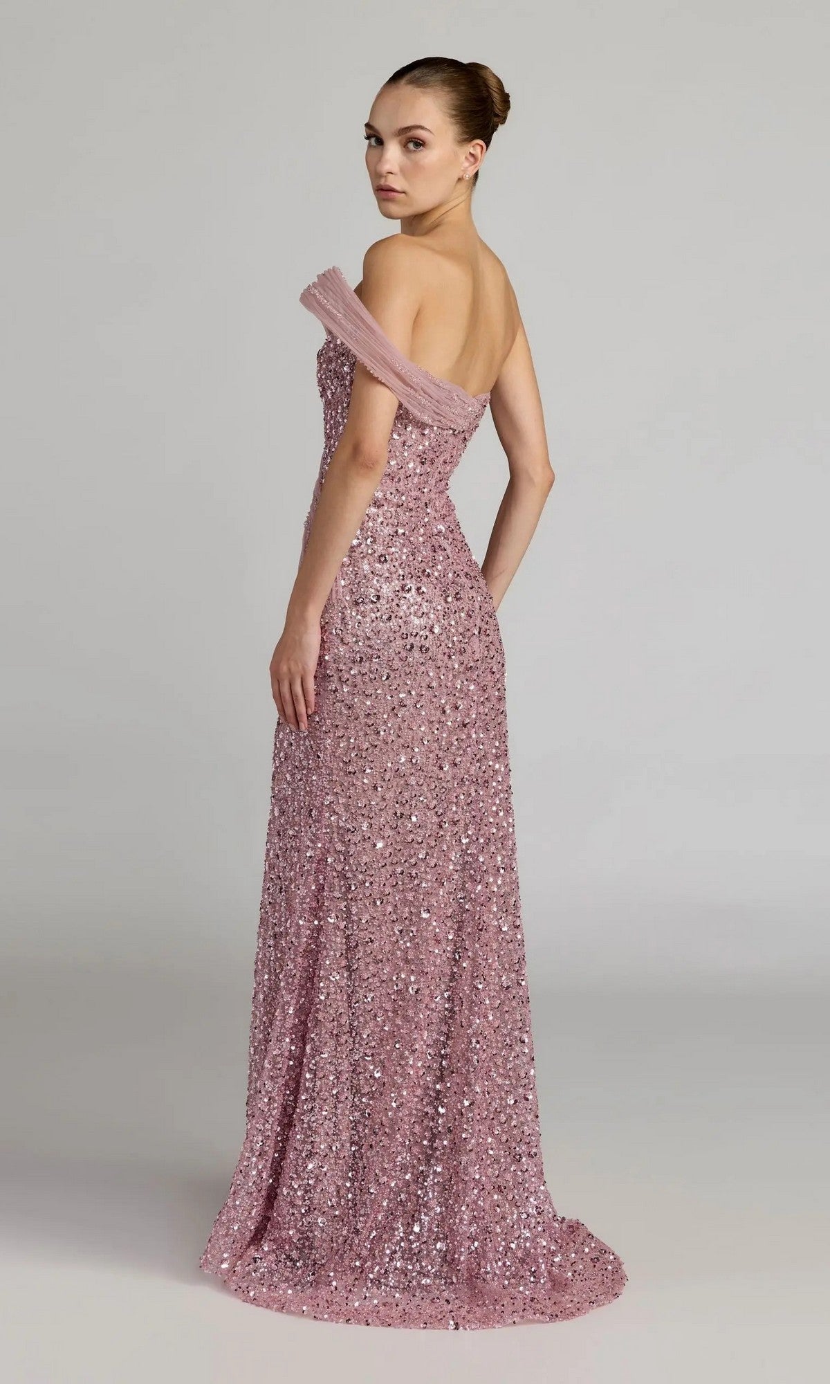 A woman poses against a plain light background, looking over her shoulder in the shimmering, off-the-shoulder, floor-length pink Long Formal Dress: Mac Duggal 37085.